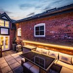 Evening garden view at The Barns, Bostock Hall Estate, showing illuminated patio areas, landscaped borders and character brick architecture